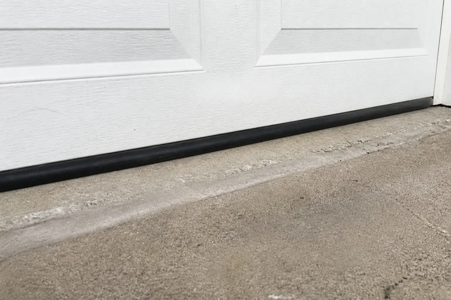 Black bottom seal along a closed white garage door at a concrete threshold. — Garage Door Bottom Seal Replacement, Kawartha Lakes