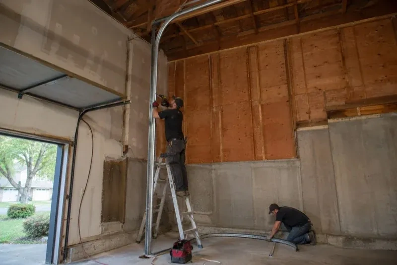 One person on a stepladder working beside a tall curved garage-door track while another kneels beside loose track sections in an unfinished garage. — High-Lift Garage Door Conversion, Kawartha Lakes
